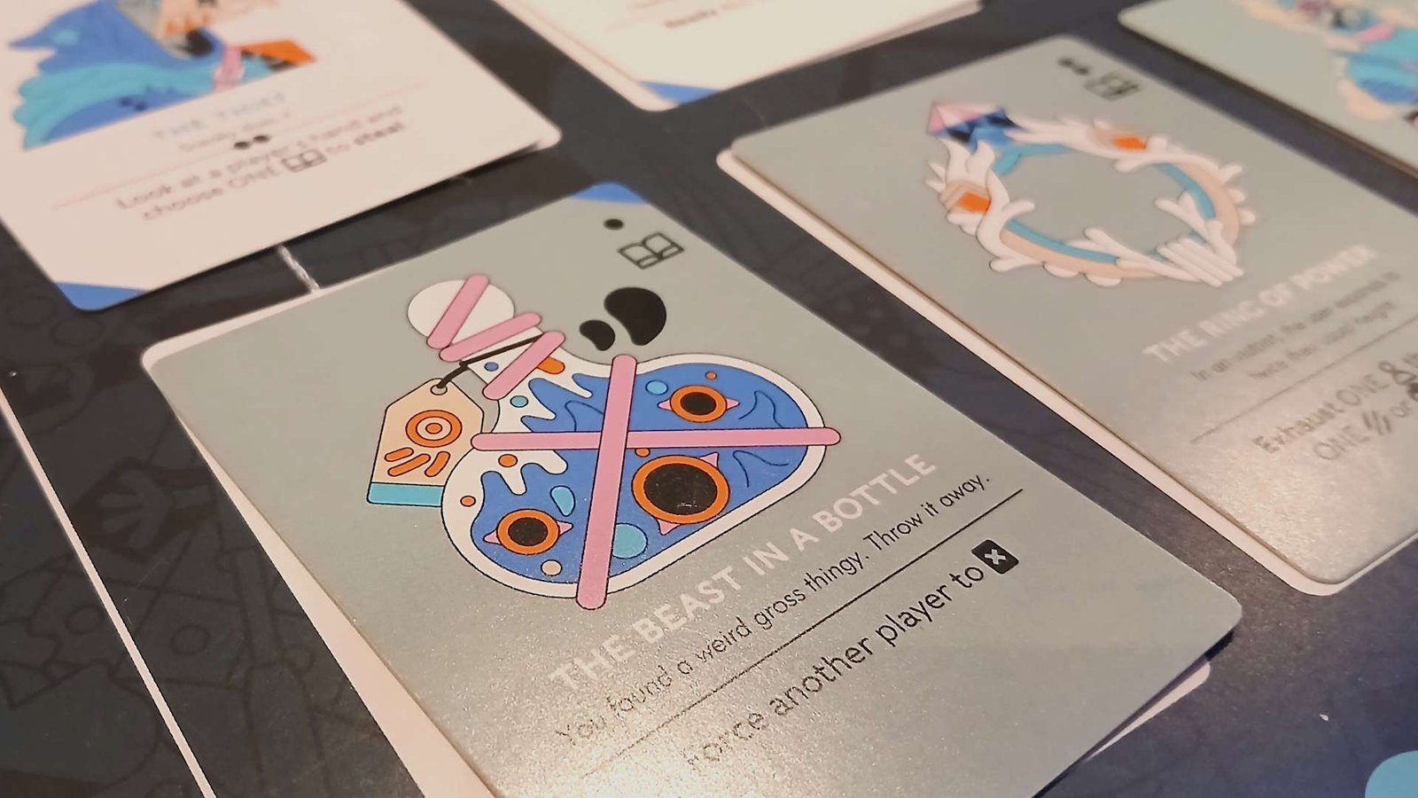 A series of cards laid out on a dark surface, with one card reading 'The Beast in a Bottle' with a cartoon bottle on it taking up much of the foreground