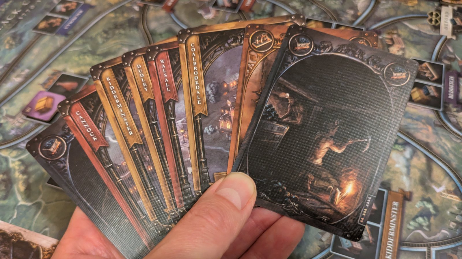 A hand holds out a spread of Brass: Birmingham cards, with the map board visible in the background
