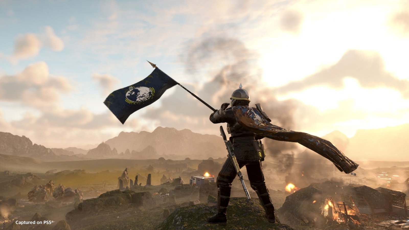 A Helldiver waves a flag while looking down at a battlefield in a screenshot from Helldivers 2's Masters of Ceremony trailer.