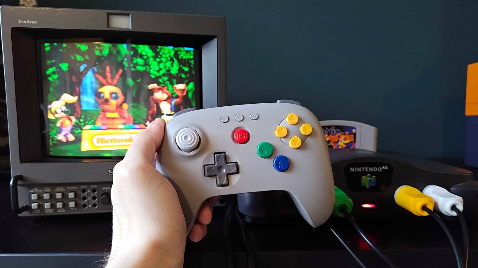 Hand holding gray 8Bitdo N64 controller in front of original console connected to Sony PVM CRT with Banjo Kazooie intro on screen.
