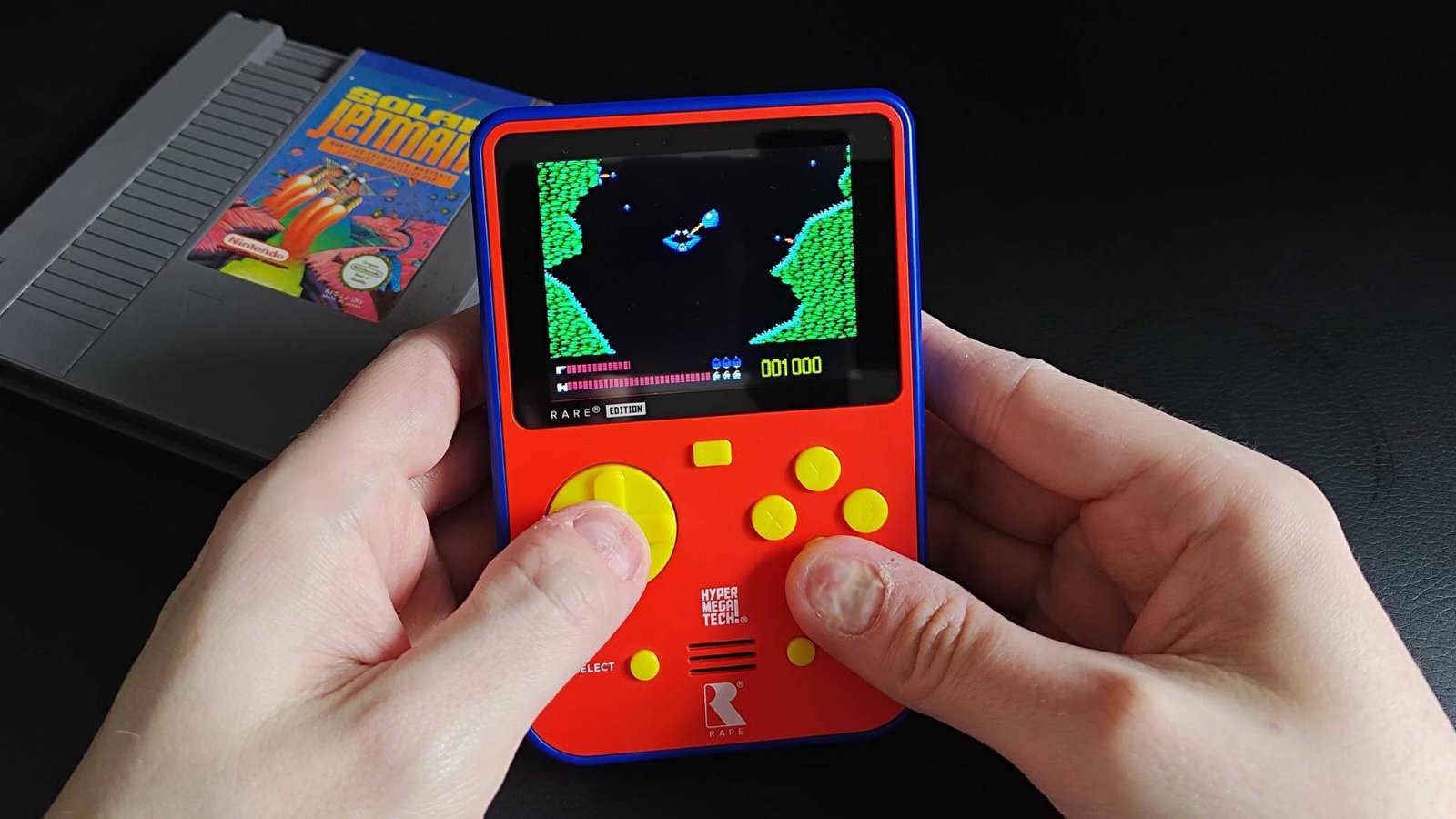 Hands using Super Pocket Rare Edition to play Solar Jetman with NES cartridge on desk in backdrop.