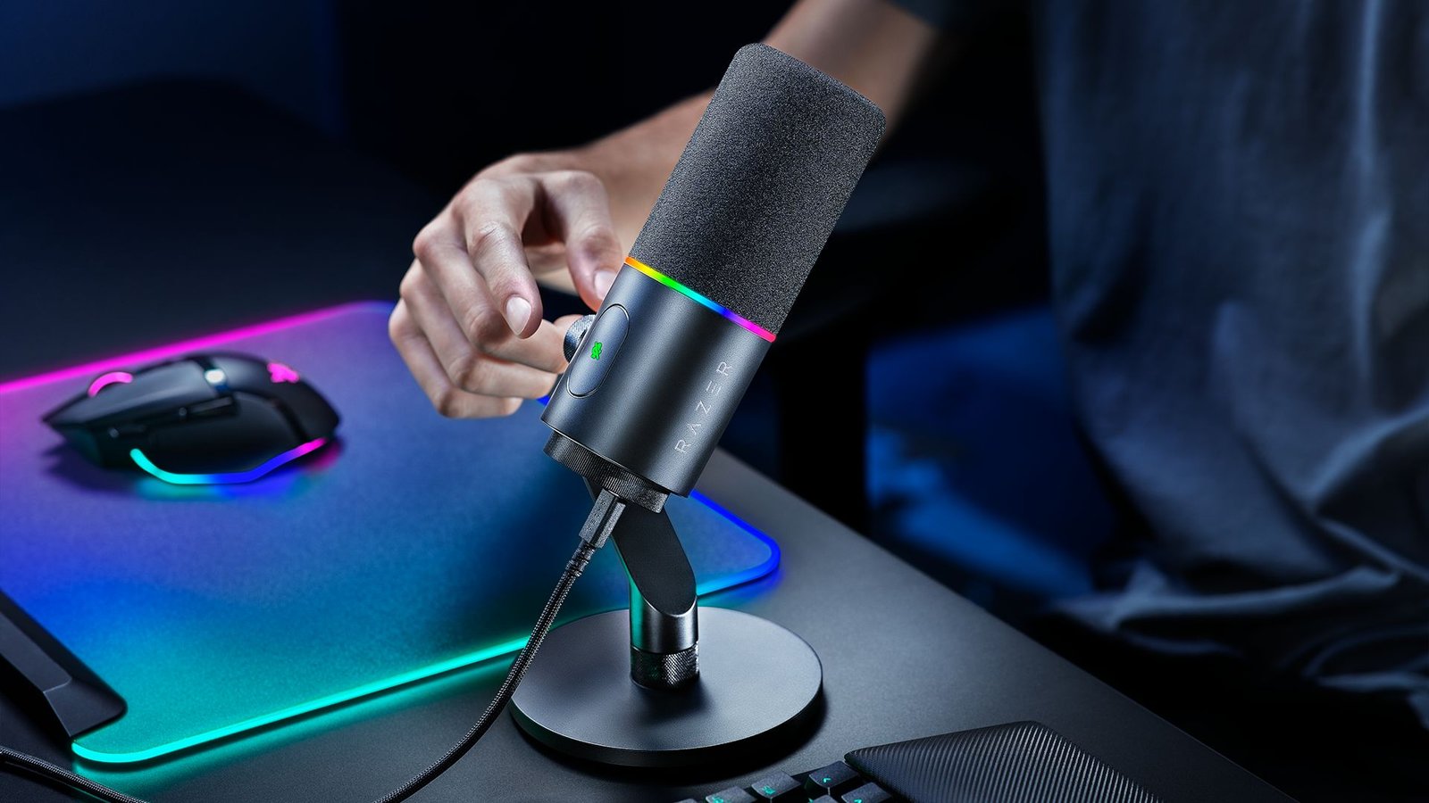 A marketing image of the Razer Seiren V3 Pro on a gaming desk with someone reaching out to press the mute button
